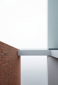 A minimalist architectural image featuring a classic red brick wall on the left, connected by a white walkway to a modern, light-filled structure on the right.